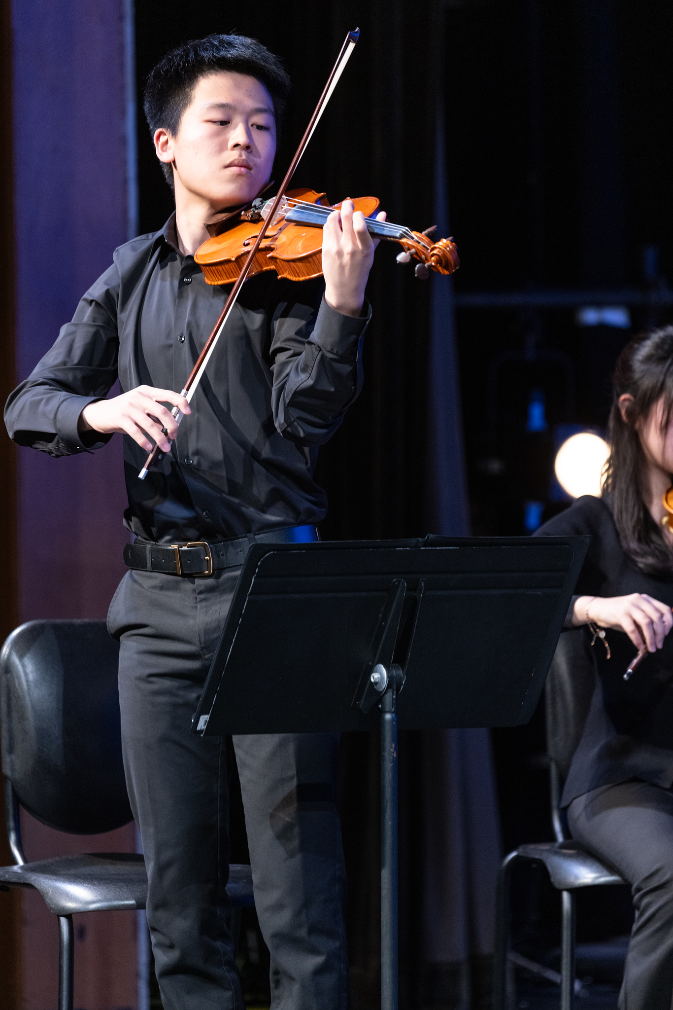 Toby playing violin on a dark stage, beside a music stand.